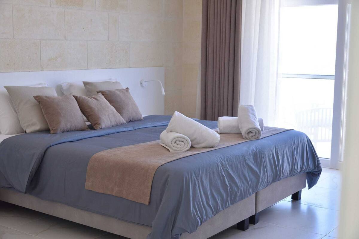Un letto rifatto con cura, con una coperta blu e beige, due asciugamani arrotolati e diversi cuscini.