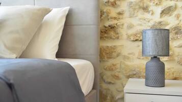 Ein Bett mit grauem Kopfteil, einem weißen Kopfkissen und einer grauen Decke. Daneben ein weißer Nachttisch mit einer grauen Lampe. Die dahinterliegende Wand weist ein Steinmuster auf.