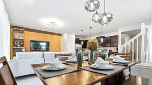 A modern living room with a wooden dining table set for four, a sofa, and a TV mounted on the wall. - Brand New, Stylish Home 5B/4, 5B, Heated Pool & BBQ (Clermont)