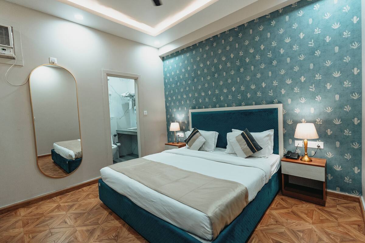 A hotel room with a large bed, a mirror, a nightstand with a lamp, and a wall with a leaf pattern.