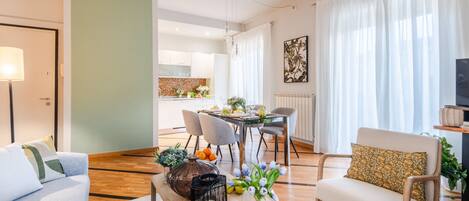 A modern living room with a sofa, a coffee table with decorative items, a dining area with chairs and a table, and a kitchen in the background.