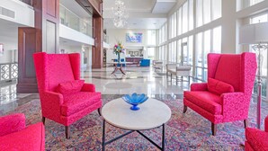 Un lobby amplio con techos altos, ventanas grandes y un área de descanso central con sillones tapizados en rojo y una mesa blanca.
