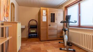 A sauna room with a wooden bench, a wall-mounted sauna heater, and a window with blinds.