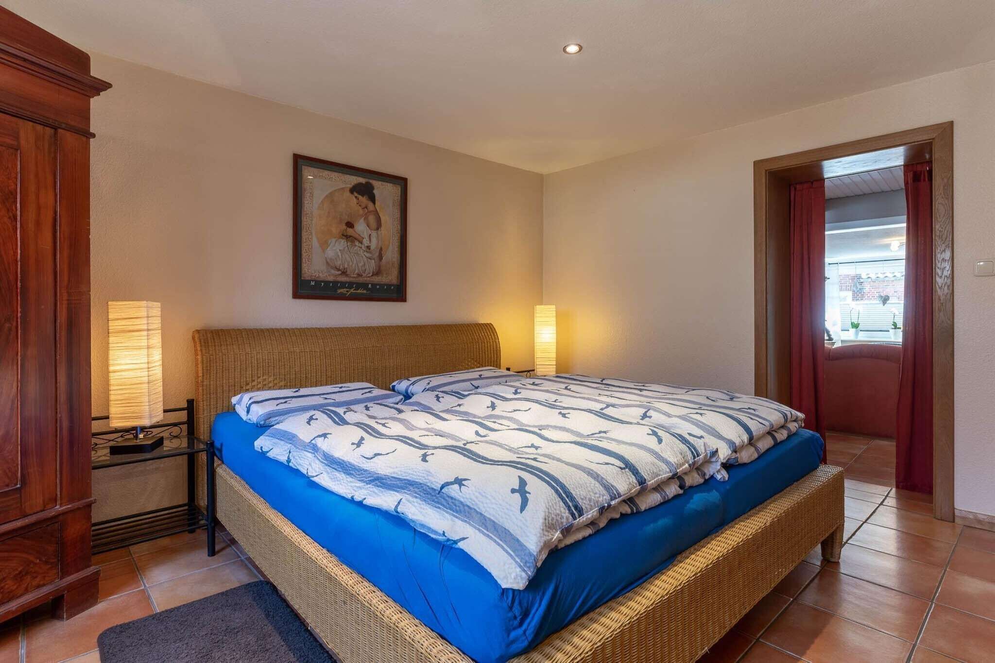 A bedroom with a bed, a nightstand, a lamp, a framed picture on the wall, and a door leading to another room.