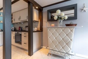 A modern kitchen with built-in appliances, a dining area with a white tufted bench, and a decorative wall-mounted shelf with flowers.