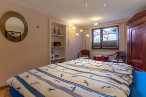A bedroom with a large bed, a chair, a wooden wardrobe, a shelf unit, and a window with blinds. - Ferienhaus in Tönning (Tönning)