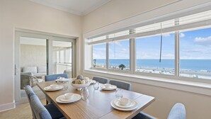 Ein Essbereich mit einem Holztisch, gedeckt für vier Personen, großen Fenstern mit Jalousien und Blick auf den Strand.