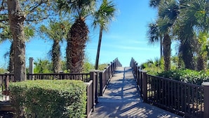 A wooden boardwalk with metal railings, surrounded by lush greenery and palm trees, leading towards a clear blue sky. - Ocean Front Villa w/ Private 4th Floor Balcony (Hilton Head Island)