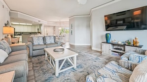 A spacious living room with a large TV, comfortable seating, a ceiling fan, and a kitchen in the background. - Ocean Front Villa w/ Private 4th Floor Balcony (Hilton Head Island)