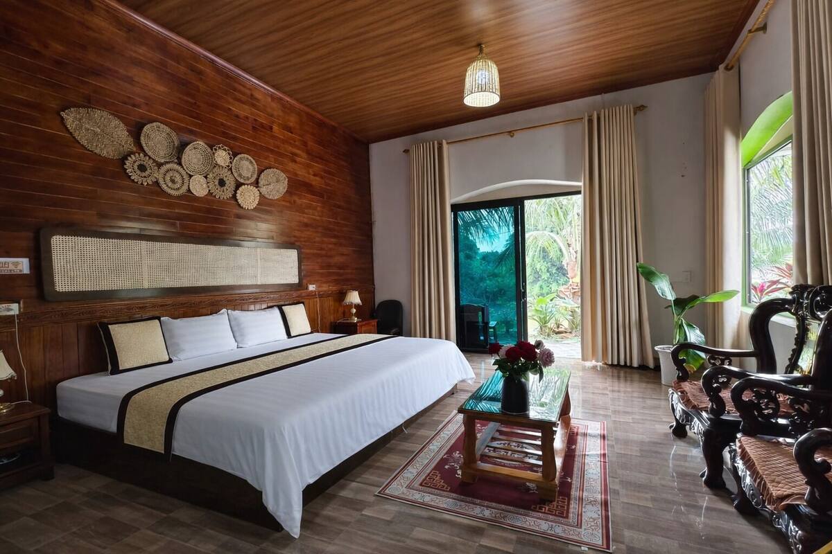Une chambre avec un plafond en bois, un grand lit, une table de chevet, une commode, une chaise, une table avec un vase, et une vue sur un jardin par une porte vitrée.