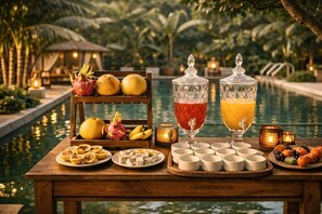 A wooden table with fruits, a fruit basket, and two glass containers with drinks, set by a poolside with palm trees and a thatched-roof structure in the background. - Hidden Valley Eco Retreat Trang An (Hoa Lu)