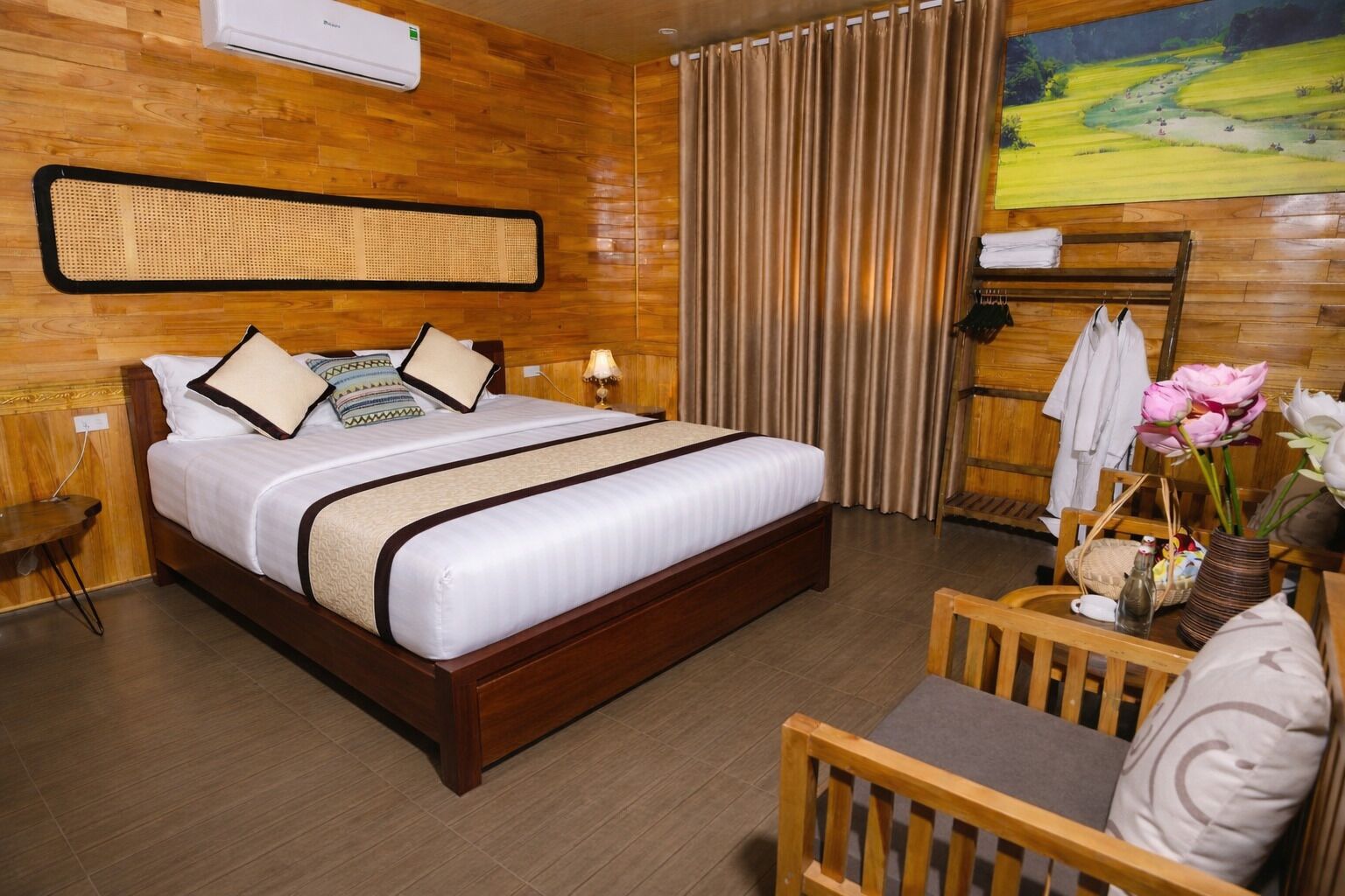 A hotel room with a wooden floor, a bed with white bedding, a rattan headboard, a wooden nightstand, a desk with a chair, and a painting of a landscape.