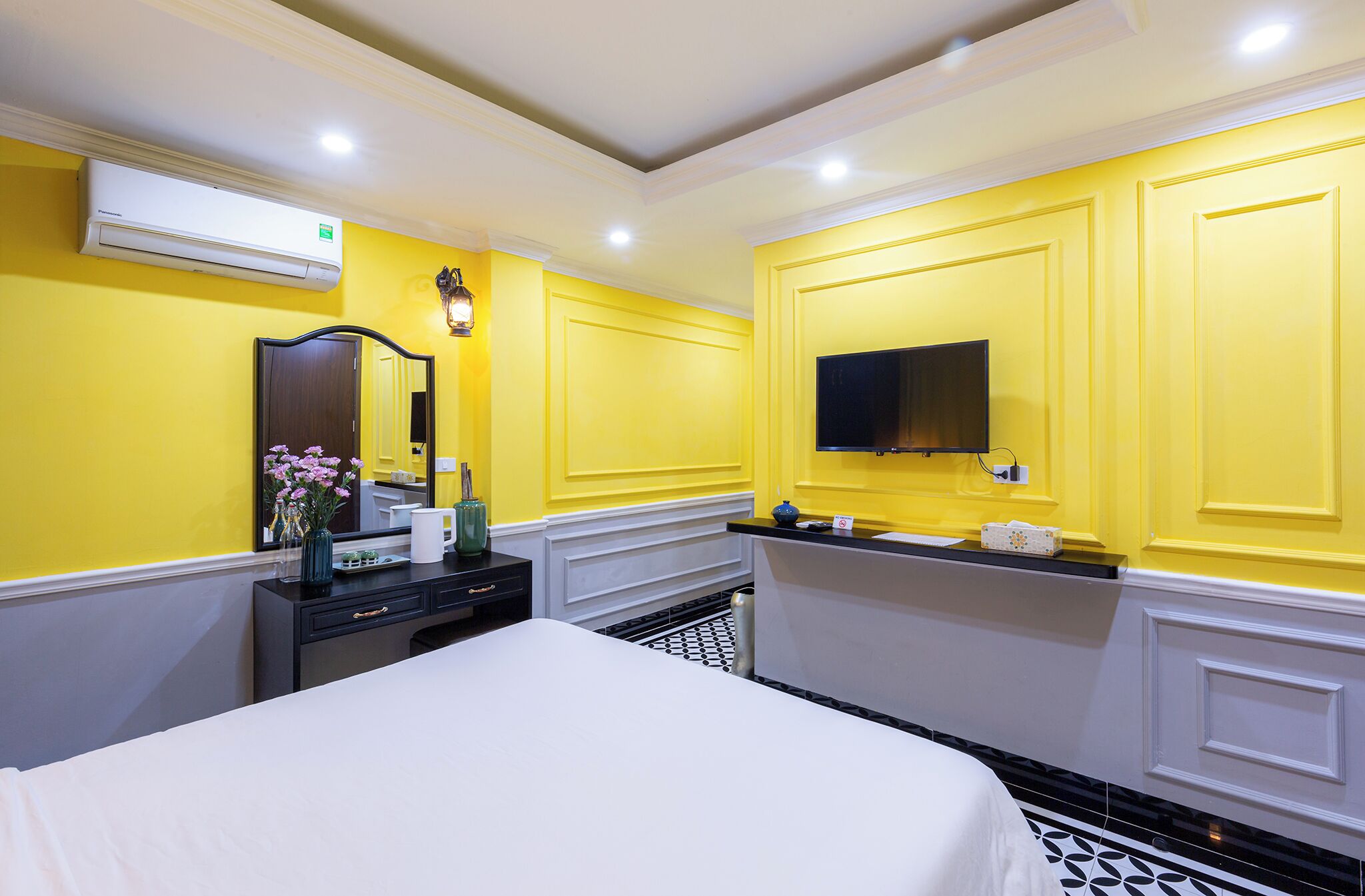 A hotel room with a bed, a dresser, a vanity with a mirror, a television mounted on the wall, and an air conditioning unit.