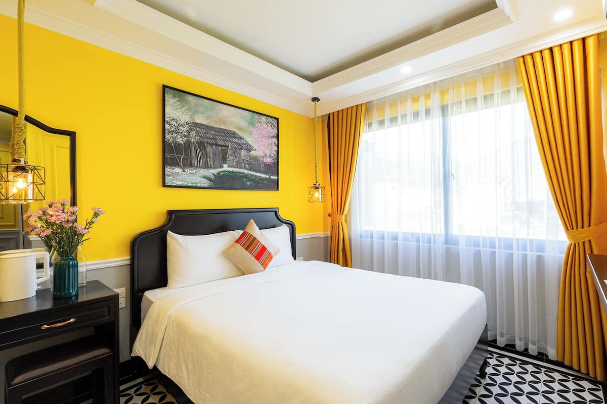 Una habitación de hotel con una cama, un cuadro, un florero con flores y una ventana con cortinas.