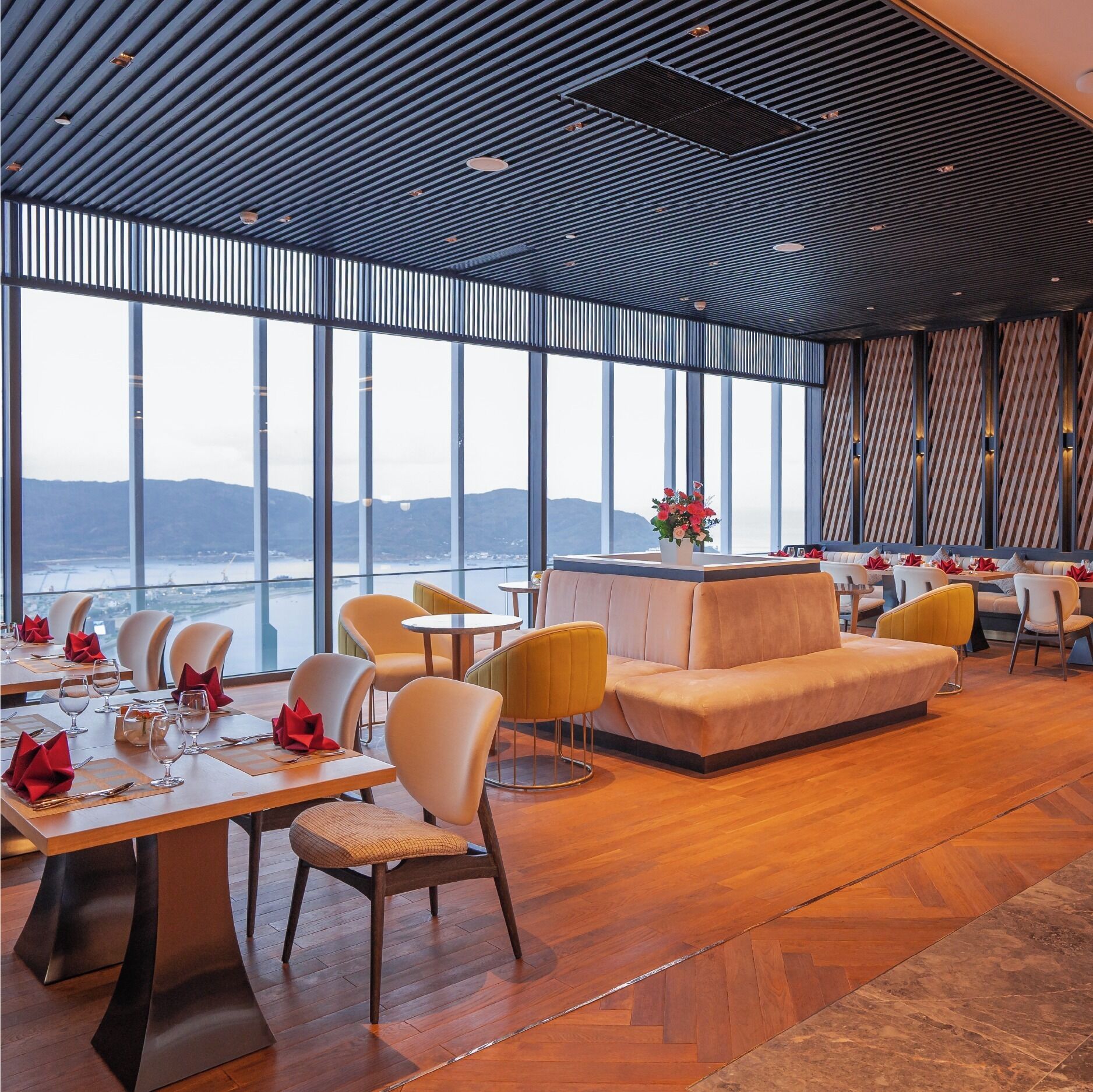 A modern dining area with a view of the sea, featuring wooden flooring, contemporary furniture, and a ceiling with a unique pattern.