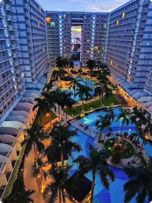 A high-rise hotel with multiple balconies, a central swimming pool surrounded by palm trees, and a well-lit outdoor area. - Shore 1 - 2BR Penthouse in MALL OF ASIA COMPLEX. (Pasay)