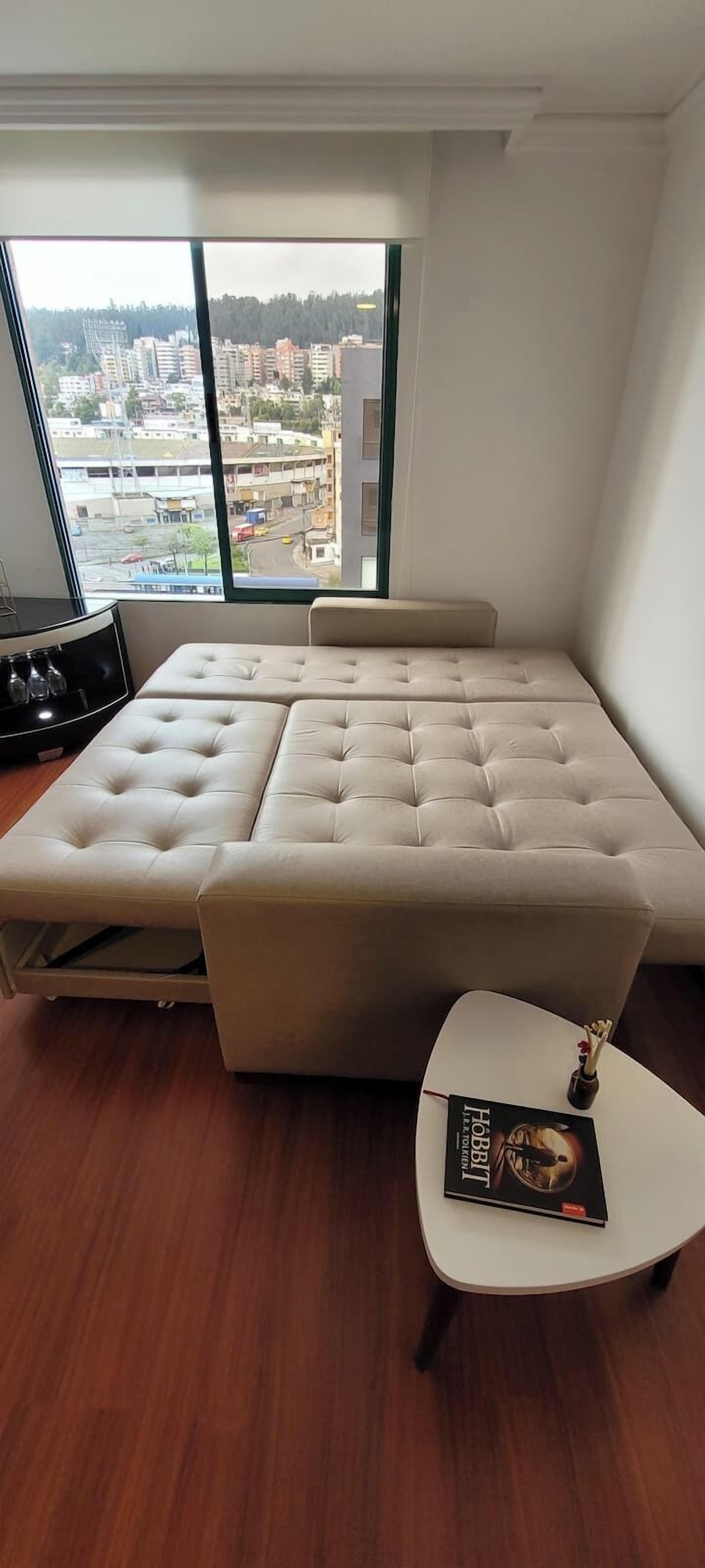 Un dormitorio moderno con un ventanal grande, un sofá cama con capitoné, una mesita de noche blanca y un libro titulado "The Hobbit".
