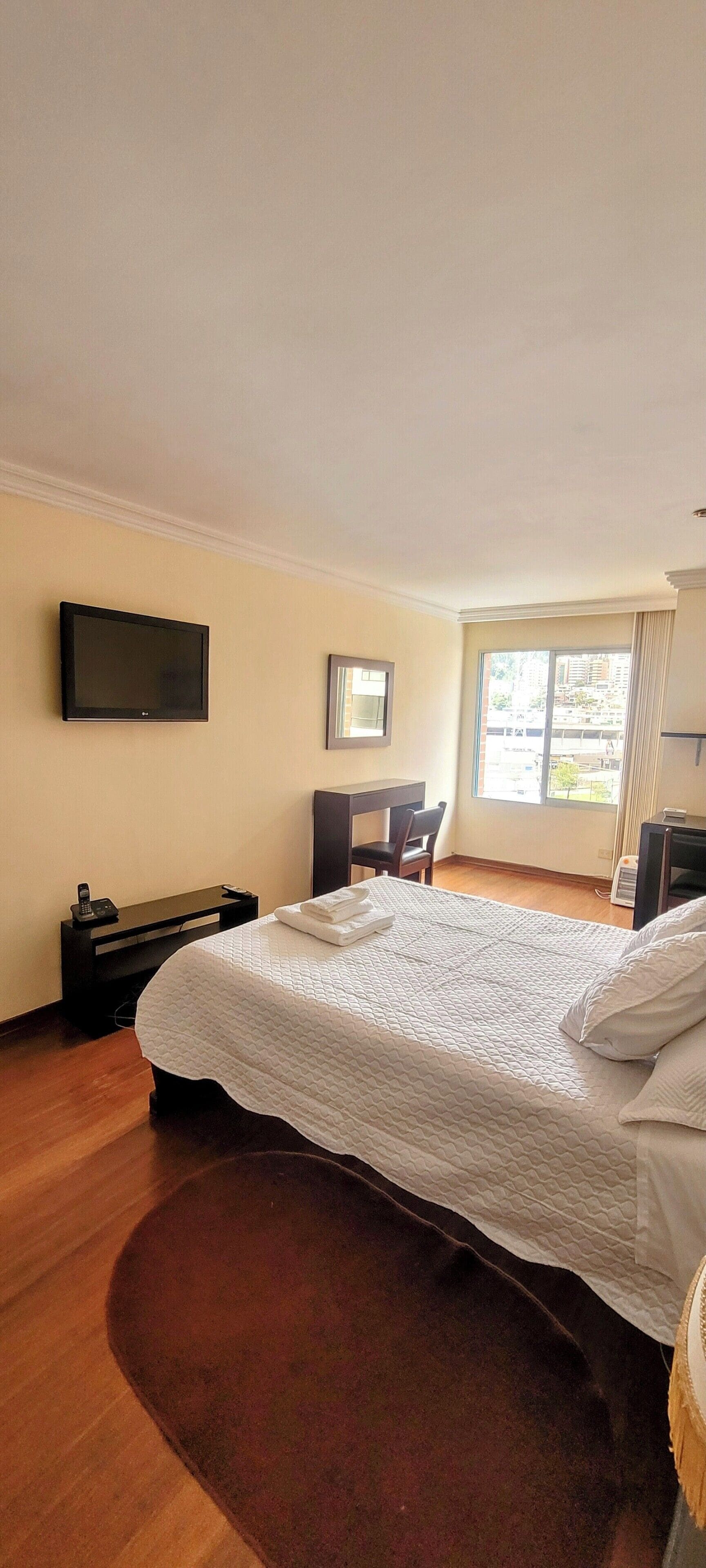 Une chambre d’hôtel avec un grand lit, un bureau, une chaise, une télévision et une fenêtre donnant sur des immeubles.