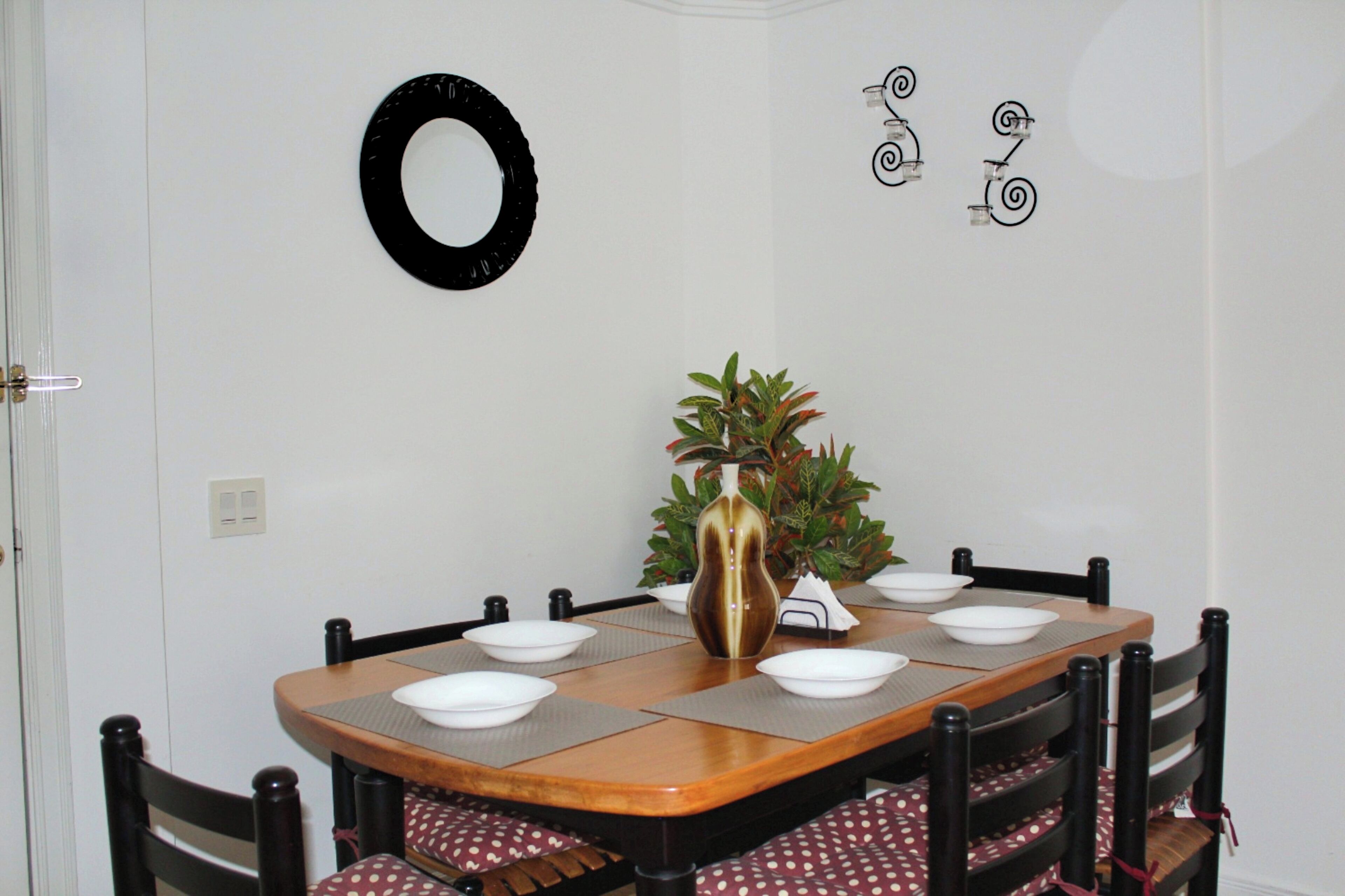 A dining table with six chairs, a vase with a plant, and wall decorations including a circular mirror and spiral designs.