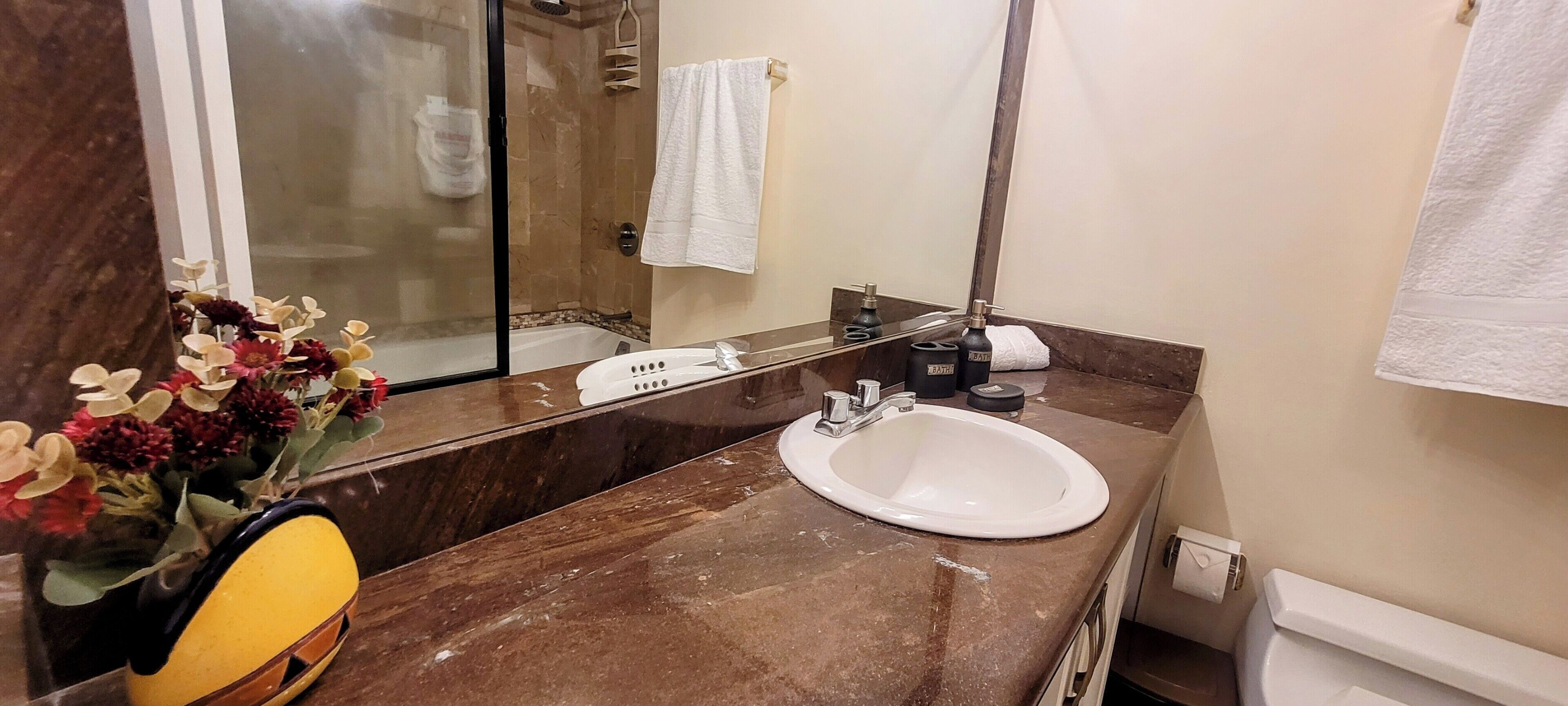 A bathroom with a marble countertop, a white sink, a mirror, a shower area, and a vase with flowers.