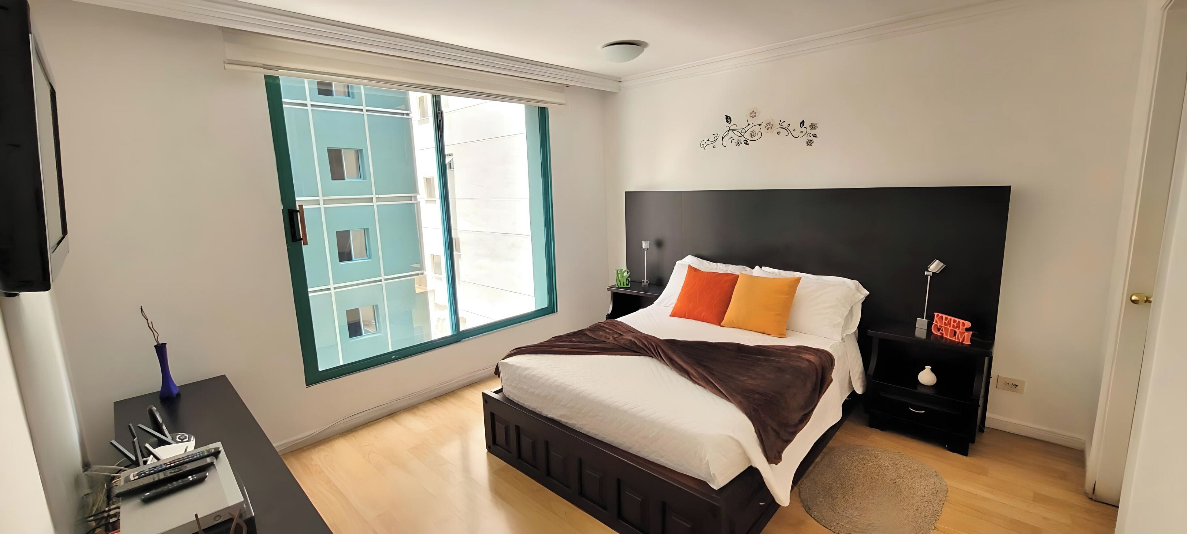 Un dormitorio con cama, mesita de noche, televisor, ventana y decoración en la pared.