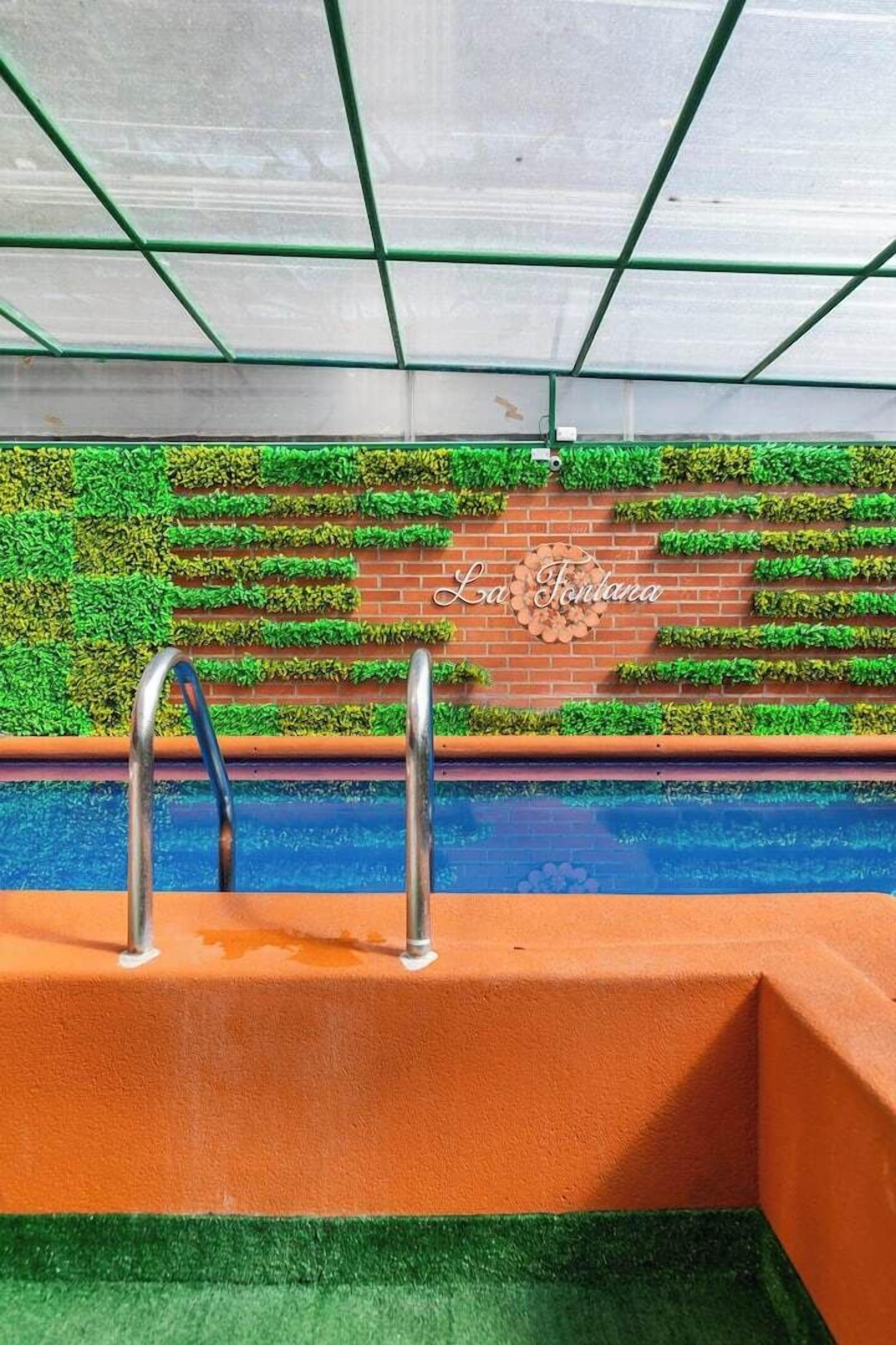 Ein Schwimmbecken mit einer Metallleiter, umgeben von einer grünen Rasenwand aus Kunststoff und einer Ziegelwand mit dem Schriftzug 'La Fontana'.