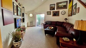 A living room with a brown leather sofa, a coffee table, and a TV. There are framed pictures on the walls and a ceiling fan. - Practical Zen: 4-bedroom 5-bed 2-bath close to everything, flexible, cookies. (Lawrence)