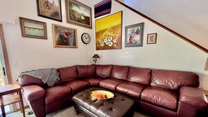 A living room with a brown leather sectional sofa, a coffee table, and various framed artworks on the walls. - Practical Zen: 4-bedroom 5-bed 2-bath close to everything, flexible, cookies. (Lawrence)