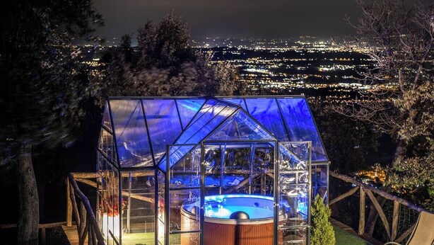 Een houten terras met een glazen hottub, omgeven door een houten hek, met op de achtergrond een stadsgezicht bij nacht.