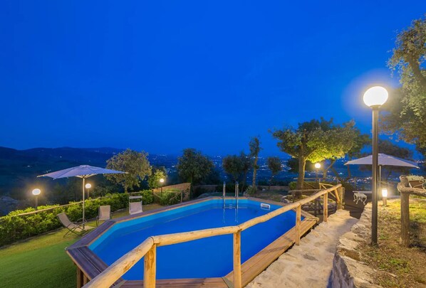 A swimming pool with a wooden deck and railings, surrounded by umbrellas and chairs, with a view of the city at dusk.