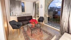 Een moderne woonkamer met een bank, twee fauteuils, een salontafel met een vaas rode bloemen en een groot raam met gordijnen.