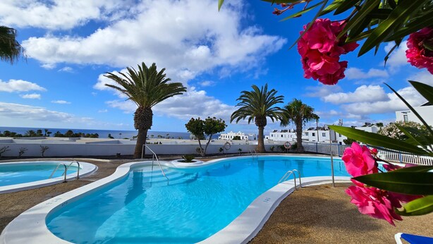 A pool area with clear blue water, surrounded by palm trees and white buildings under a partly cloudy sky. - Panoramic sea views, sunny, private balcony, refurbished on Atalaya Complex (Tías)