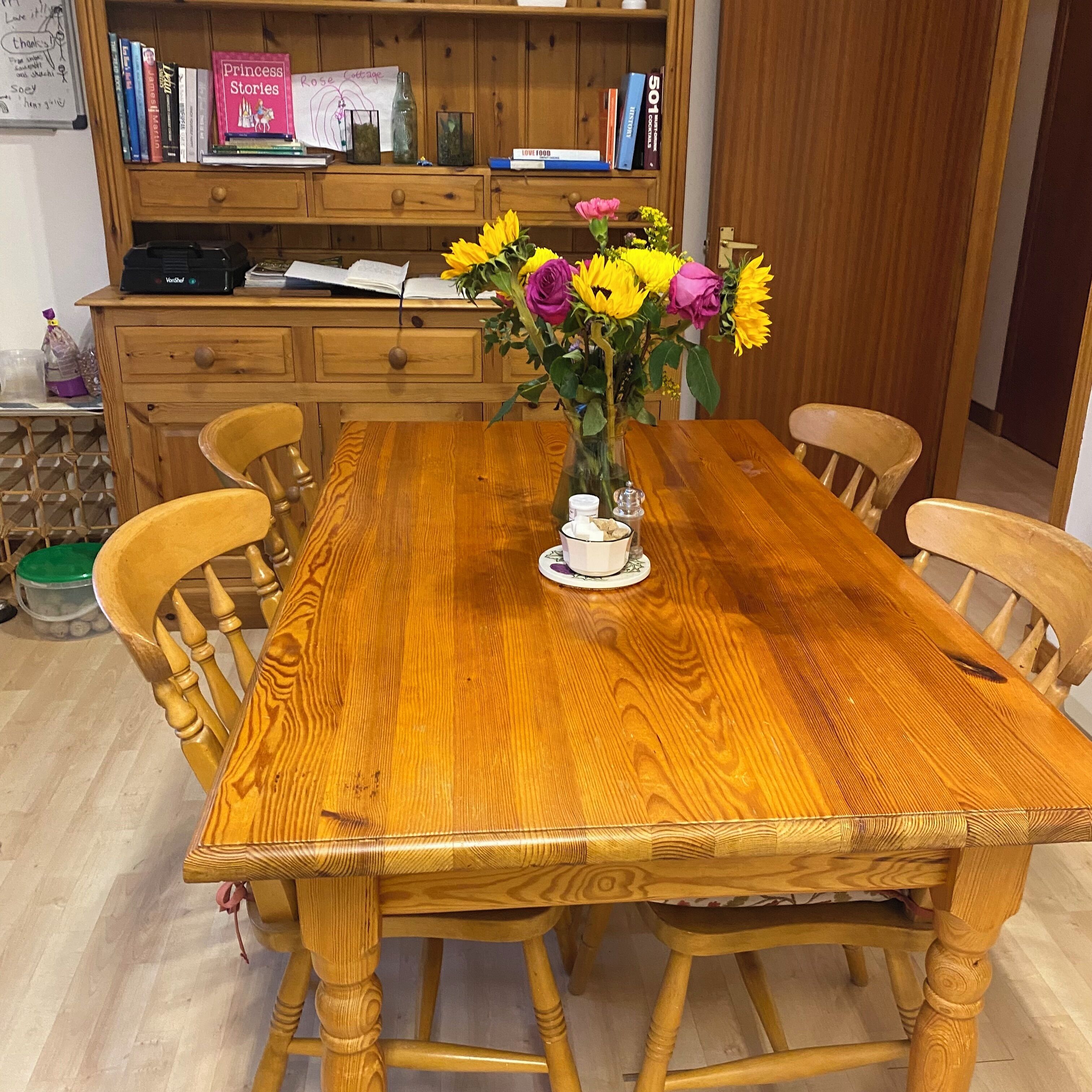 Une table à manger en bois, avec des chaises, un vase rempli de fleurs et une bibliothèque contenant des livres et des objets décoratifs.