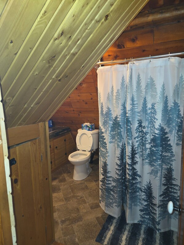 Un baño con techo de madera, inodoro, cortina de ducha con estampado de bosque y un mueble pequeño.