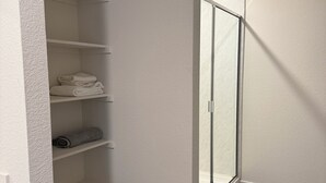 Eine Raumecke mit weißer Wand mit Struktur, Holzboden, einem weißen Mülleimer und einem Kleiderschrank mit offenen Regalen.