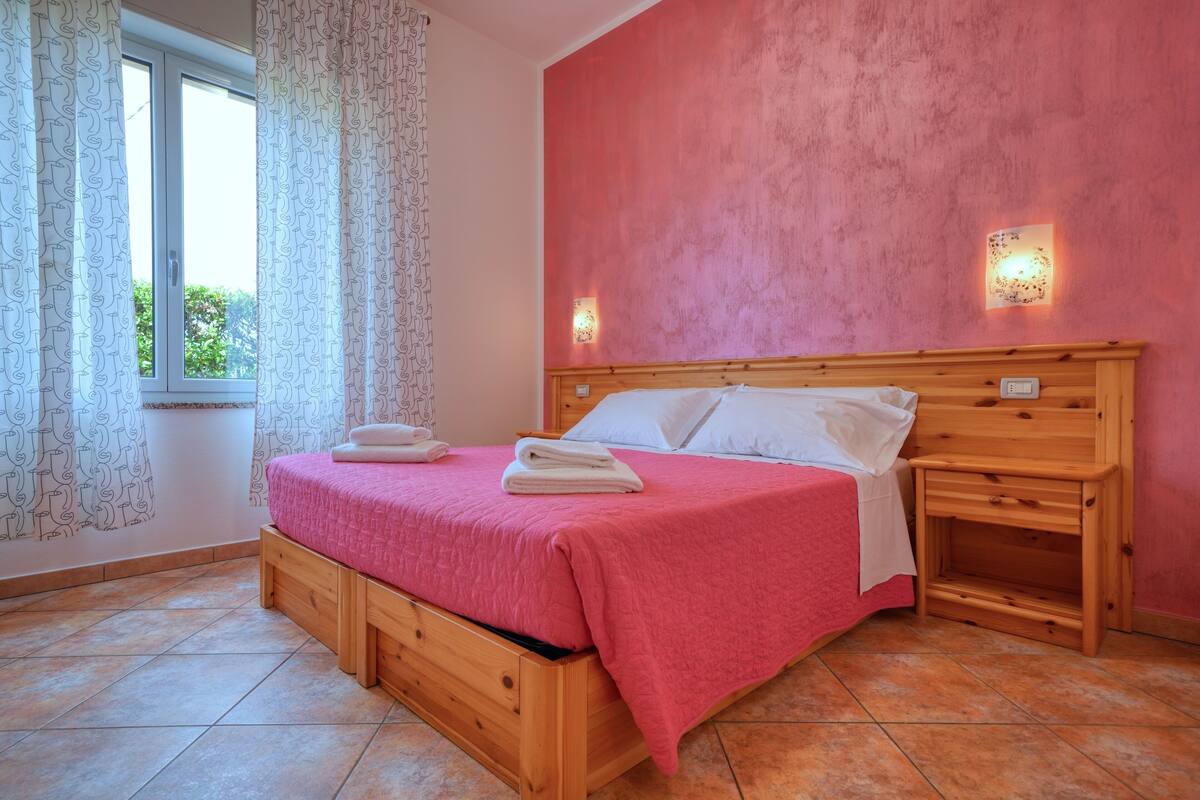 A bedroom with a wooden bed, white pillows, a pink bedspread, a nightstand, and a window with curtains.