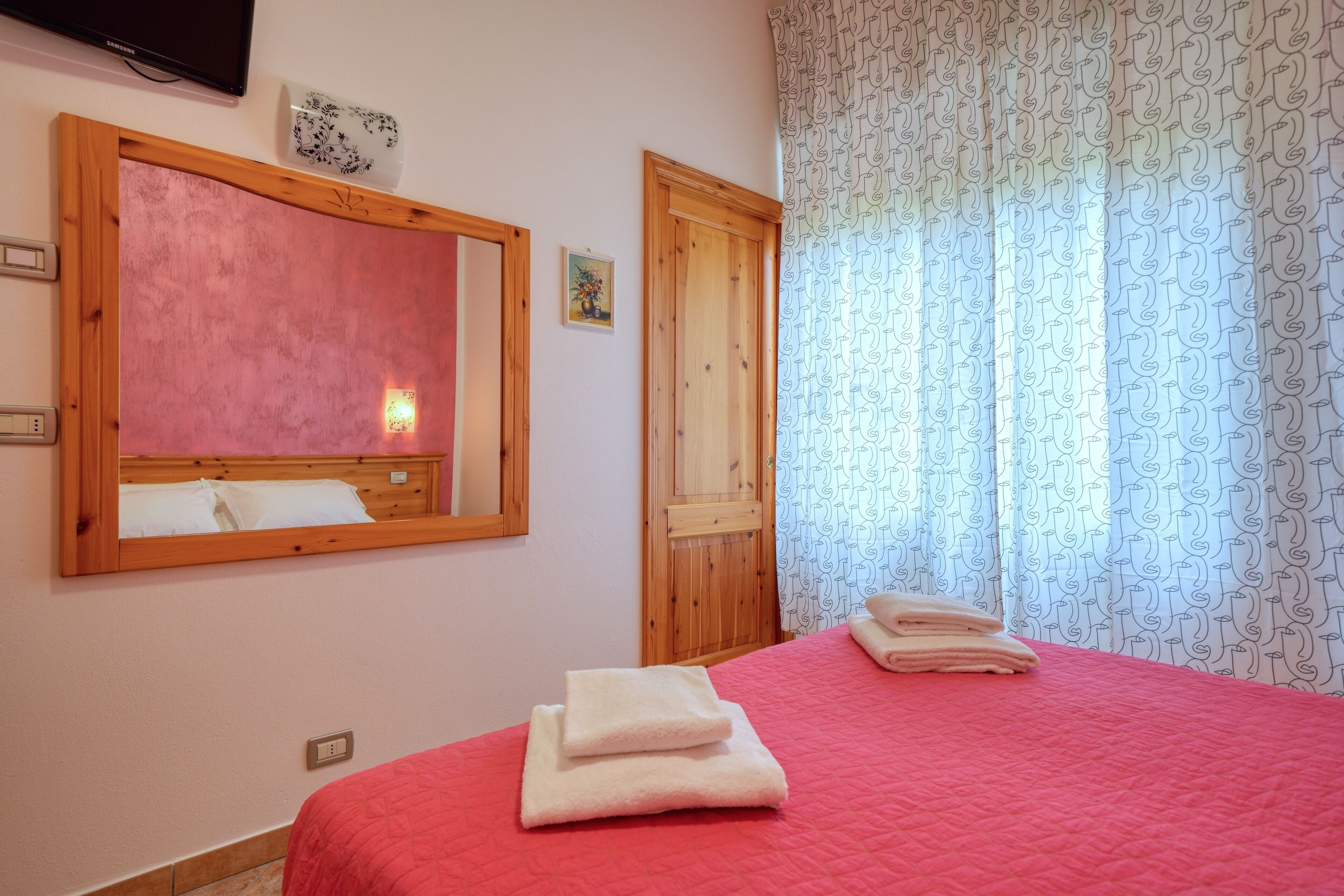 Un dormitorio con colcha roja, cabecera de madera, un espejo grande y una puerta con marco de madera.