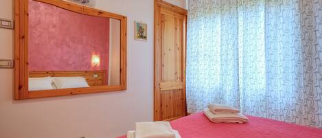 Un dormitorio con colcha roja, cabecera de madera, un espejo grande y una puerta con marco de madera.