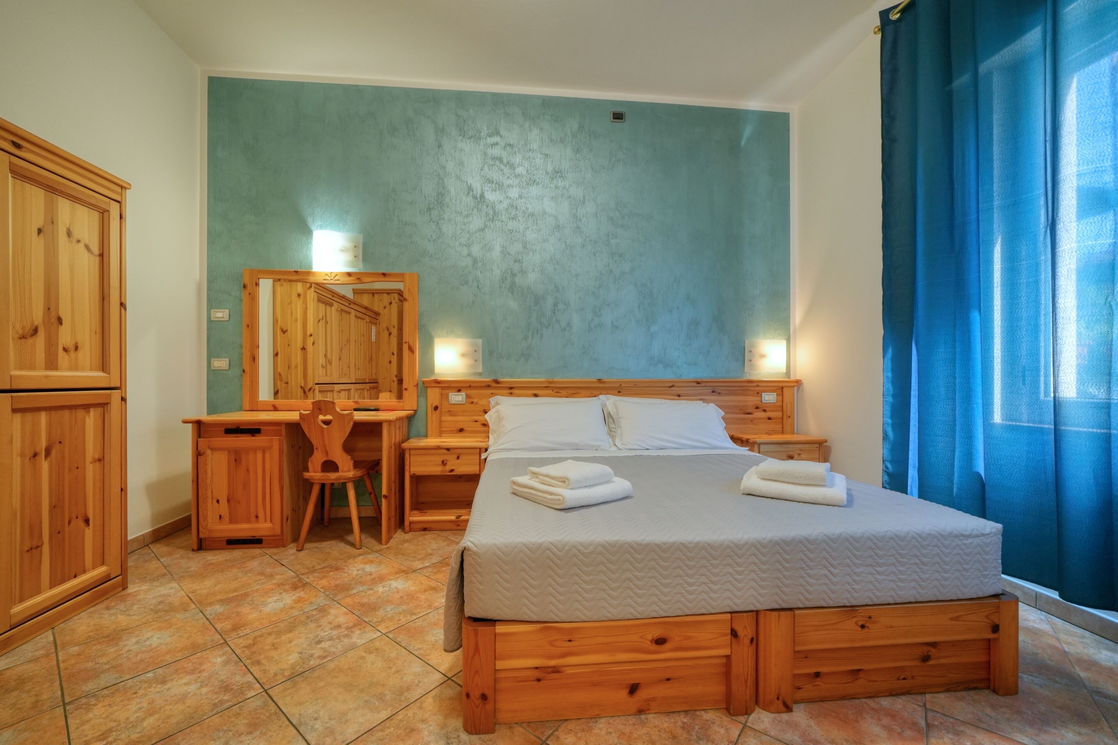Una cama de madera con colchón gris, una mesita de noche de madera, un escritorio de madera con silla, un armario de madera y una cortina azul.