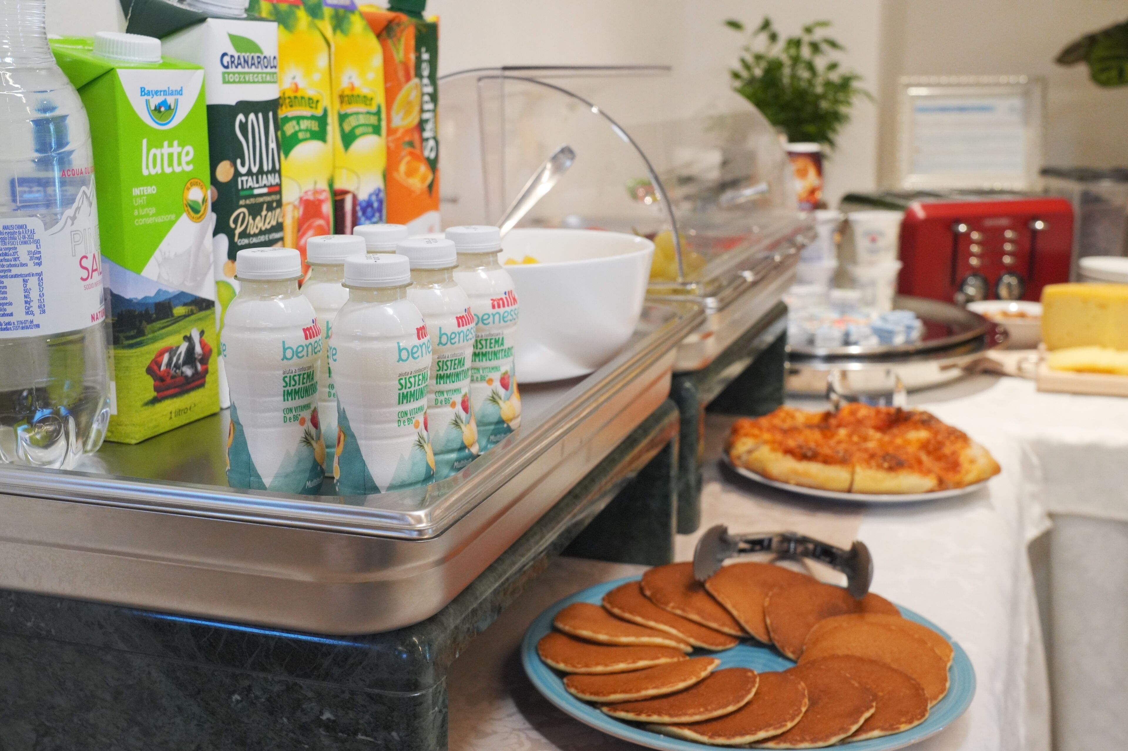 Zona de buffet con diferentes alimentos y bebidas, incluyendo una fuente de tortitas, un bol de cereales y varias cajas de leche y zumo.