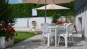 Un patio con una mesa y sillas bajo un paraguas grande, rodeado de plantas en macetas y una pared blanca.
