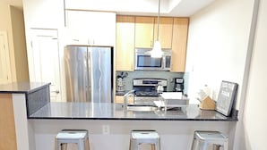 A modern kitchen with stainless steel appliances, wooden cabinets, and a black countertop. There are three metal stools and a patterned rug on the wooden floor.