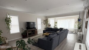 A living room with a large grey sectional sofa, a flat-screen TV, and various potted plants. - Cozy 3 bedroom home by the beach with designated work space (Long Beach)