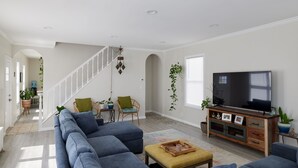 A spacious living room with a large sofa, a coffee table, and a TV on a wooden cabinet. There is a staircase leading to another room, and a dining area with chairs and a table. - Cozy 3 bedroom home by the beach with designated work space (Long Beach)