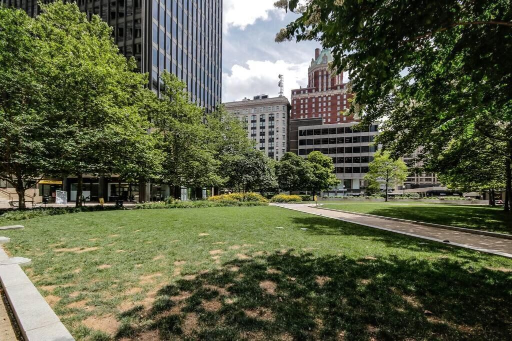 A park with a paved walkway, surrounded by trees and buildings.