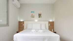 A neatly made bed with white linens and towels, a wooden headboard, bedside tables with lamps, and two framed pictures on the wall. - WI-FI 500MB | 50” Smart TV | Swimming Pool #CA67 (Florianópolis)