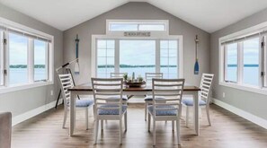 Zona pranzo con tavolo e quattro sedie, ampie finestre vista sull'acqua e un cartello sopra le finestre con scritto "Beach Cottage".