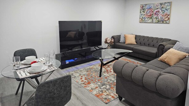 A modern living room with a sofa, a glass coffee table, a dining table set with plates and glasses, and a flat-screen TV. - Contemporary Two Bedroom Basement Suite. (Edmonton)