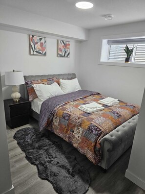 Ein Schlafzimmer mit einem grau bezogenen Bett, einer gemusterten Tagesdecke, einem dunklen Teppich, einem Nachttisch mit Lampe und zwei Wandbildern.