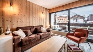 Ein modernes Wohnzimmer mit einem braunen Sofa, einem Holztisch und einem roten Sessel. Ein großes Fenster bietet Blick auf einen verschneiten Berg und Gebäude.